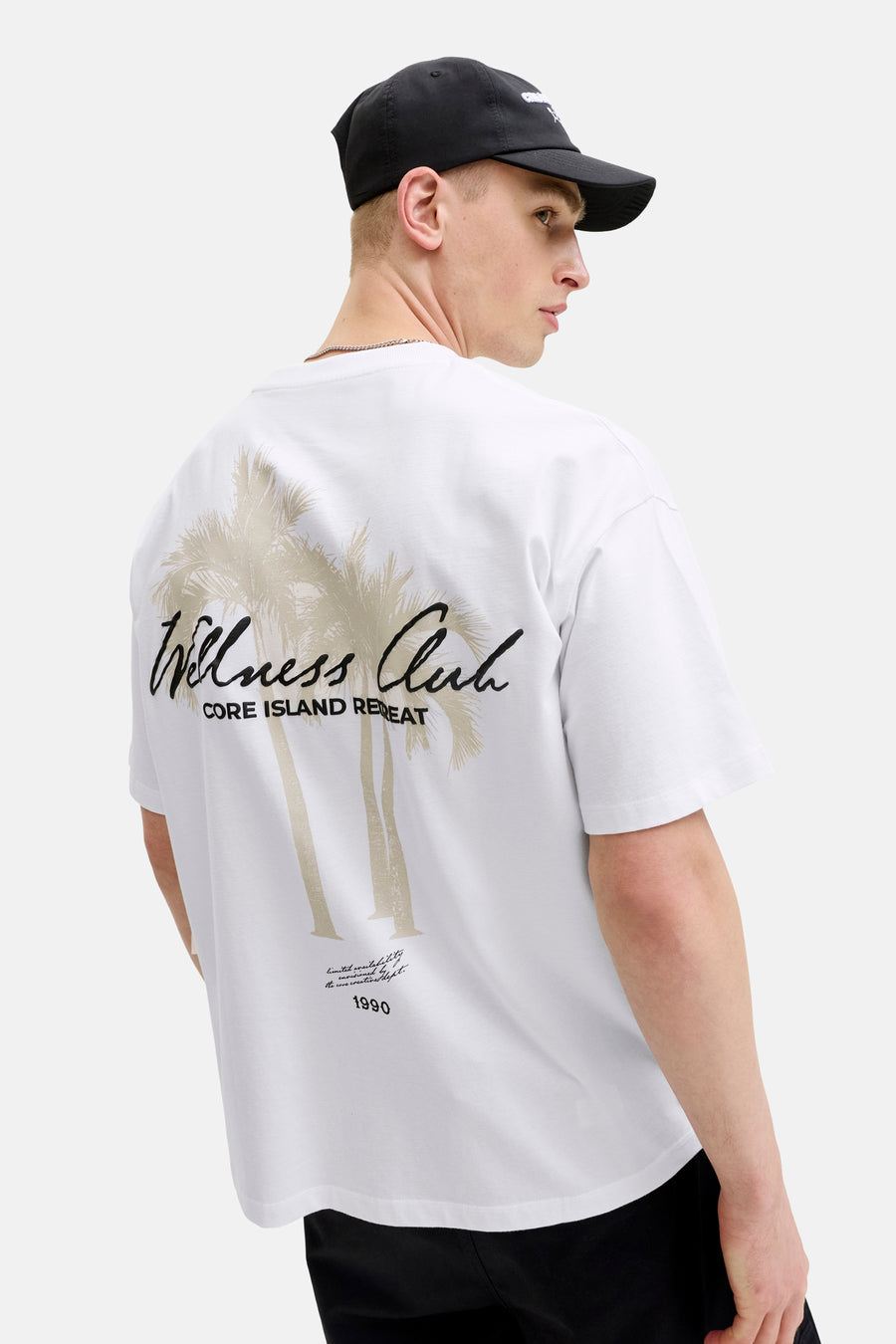 Wit T-shirt met korte mouwen van CORE BY JACK & JONES, met 'Wellness Club CORE ISLAND RETREAT' en palmbomen op de rug, gedragen door een man.