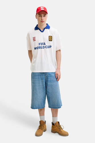 Witte polo van ORIGINALS BY JACK & JONES met blauwe kraag, FIFA World Cup- en 'CAN MEX USA'-opdruk, gecombineerd met denim shorts en rode pet.