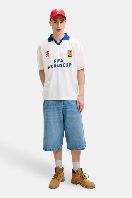 Witte polo van ORIGINALS BY JACK & JONES met blauwe kraag, FIFA World Cup- en 'CAN MEX USA'-opdruk, gecombineerd met denim shorts en rode pet.