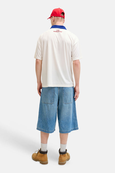 Polo blanc ORIGINALS BY JACK & JONES avec col bleu et inscription rouge 'FIFA WORLD CUP™' dans le dos, porté avec une casquette rouge et un short en jean.