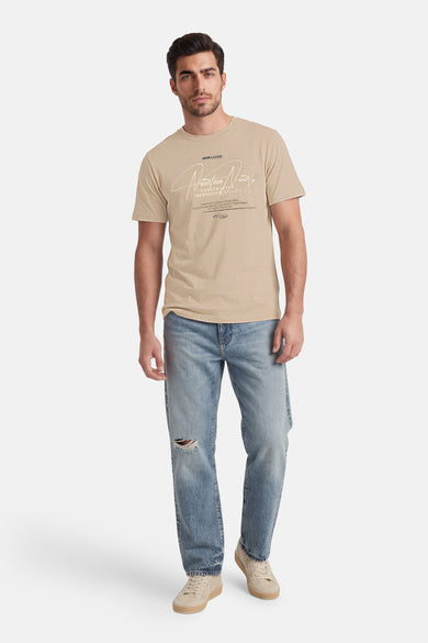 T-shirt beige à manches courtes de CORE BY JACK & JONES, avec imprimé texte blanc, porté avec un jean déchiré et des baskets beiges.