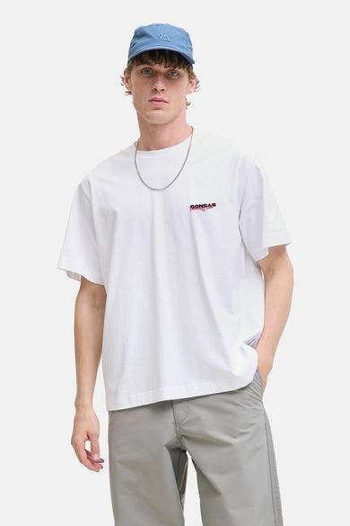 T-shirt blanc à manches courtes d'ORIGINALS BY JACK & JONES, avec un petit imprimé texte et porté avec un chapeau bleu et une chaîne argentée.