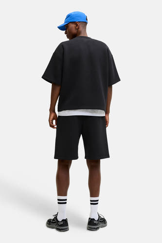 Zwart oversize sweater met ronde hals van CORE BY JACK & JONES, achterkant weergegeven, gedragen met witte ondertrui, zwarte shorts en blauwe pet.