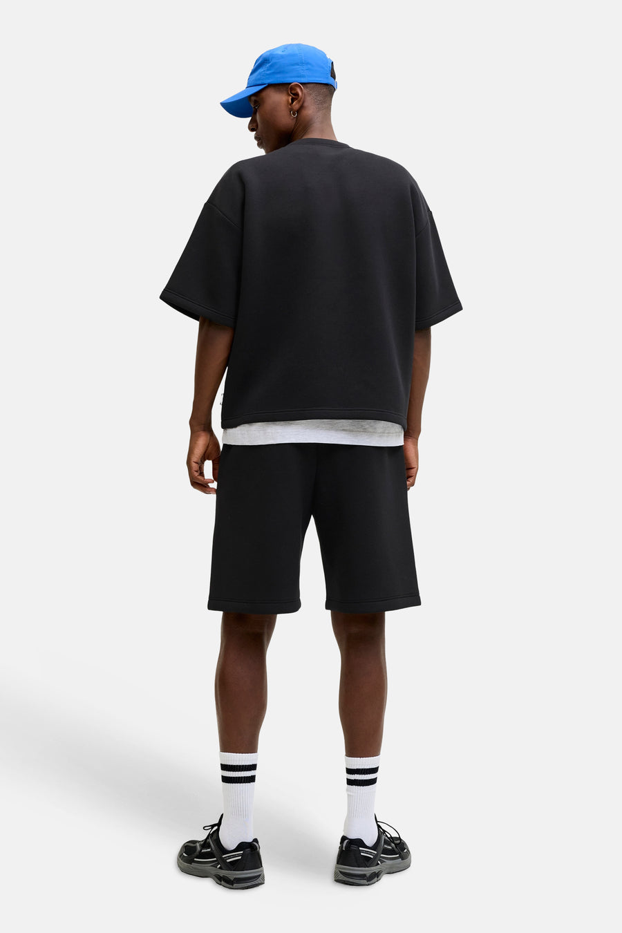 Zwart oversize sweater met ronde hals van CORE BY JACK & JONES, achterkant weergegeven, gedragen met witte ondertrui, zwarte shorts en blauwe pet.