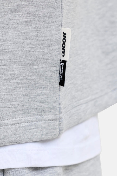 Grijze ronde hals sweater van CORE BY JACK & JONES, met zachte melange stof en witte label, gedragen over een witte t-shirt, close-up detail.