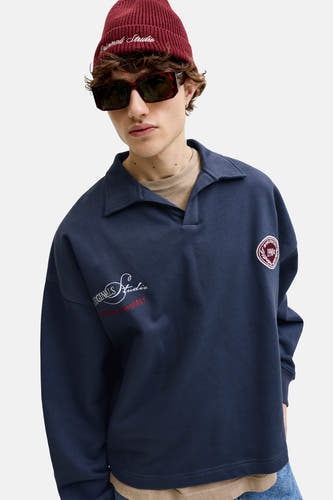 Donkerblauwe sweater met kraag van ORIGINALS BY JACK & JONES, met geborduurde witte en rode logo's op de mouw en borst, gedragen door een man met rode muts en zonnebril.