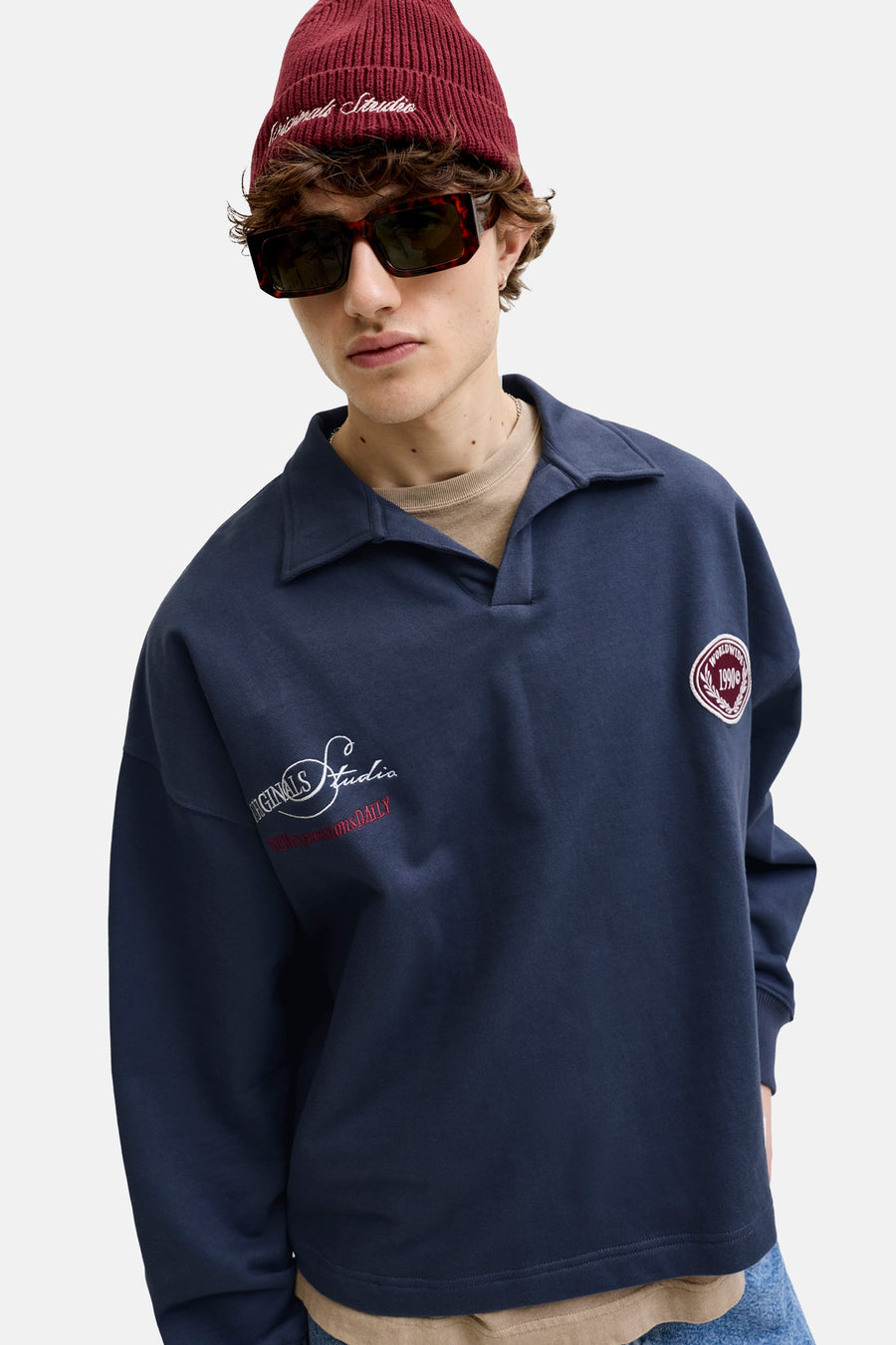 Donkerblauwe sweater met kraag van ORIGINALS BY JACK & JONES, met geborduurde witte en rode logo's op de mouw en borst, gedragen door een man met rode muts en zonnebril.
