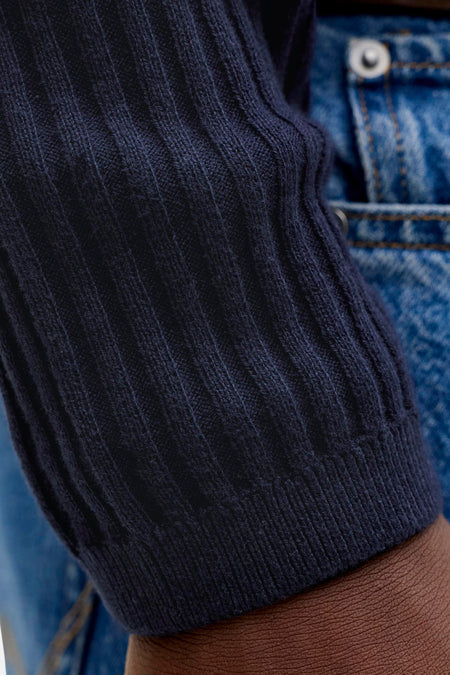 Pull en tricot côtelé bleu foncé de PREMIUM by JACK & JONES, gros plan sur la manche par-dessus un jean et peau visible.