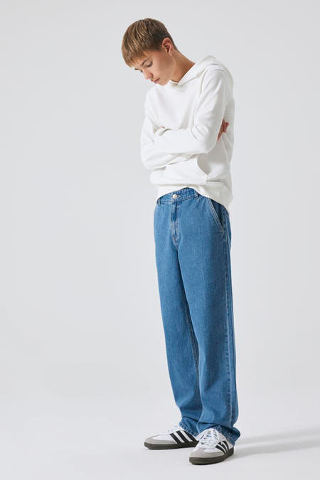 Jean large modèle de LMTD, en denim bleu moyen, porté avec une hoodie blanche et des baskets grises.