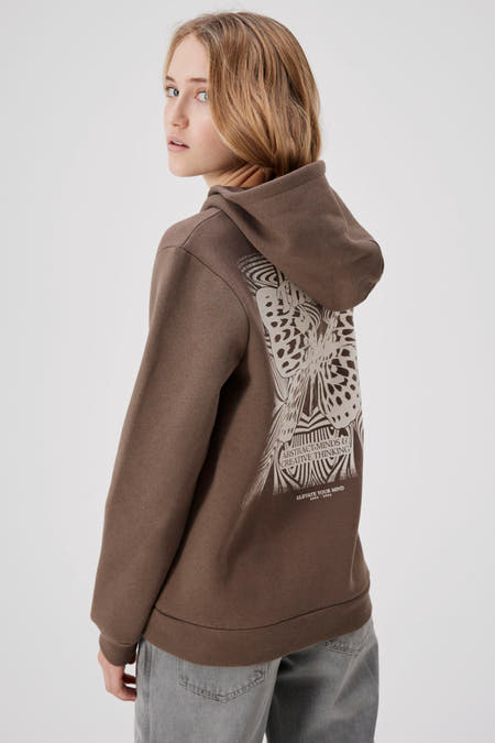 Bruine sweater met kap van LMTD, met grafische print op de rug, gedragen met een grijze jeans.
