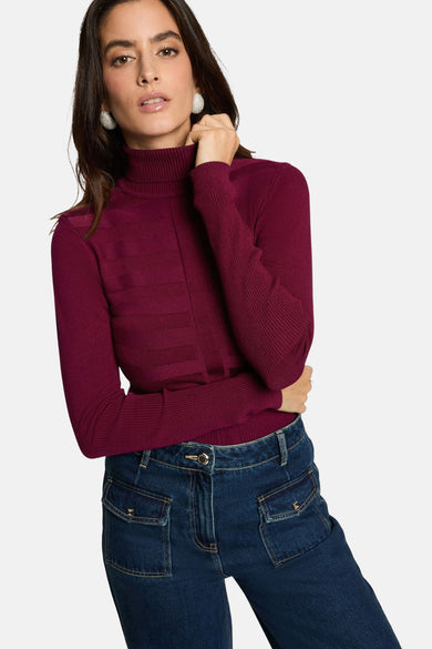 Vrouw met donkerbruin haar, bordeauxrode coltrui en donkerblauwe jeans met zakken, lichtgrijze achtergrond.