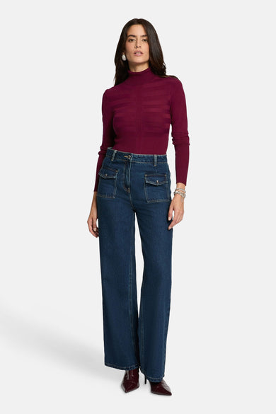 Vrouw in bordeauxrode coltrui en blauwe jeans met wijde pijpen, staand.