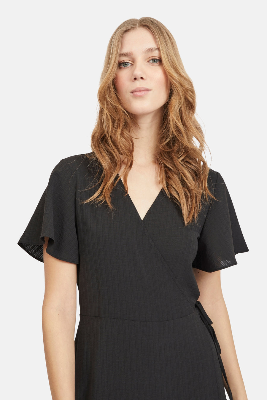 Vrouw in zwarte wikkeljurk met golvend blond haar, witte achtergrond.