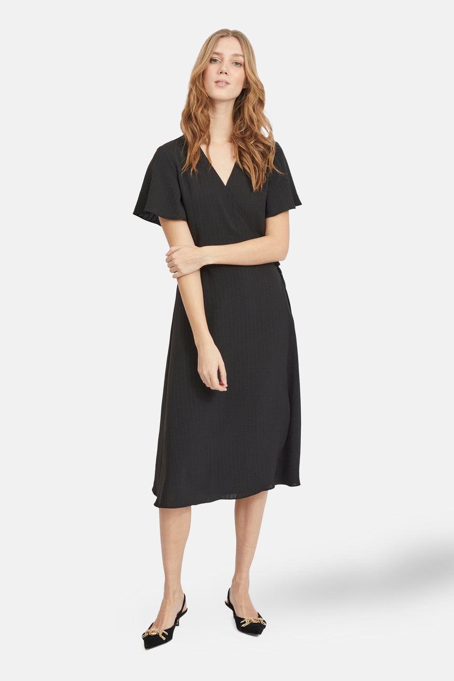 Vrouw in zwarte wikkeljurk met korte mouwen en zwarte schoenen.