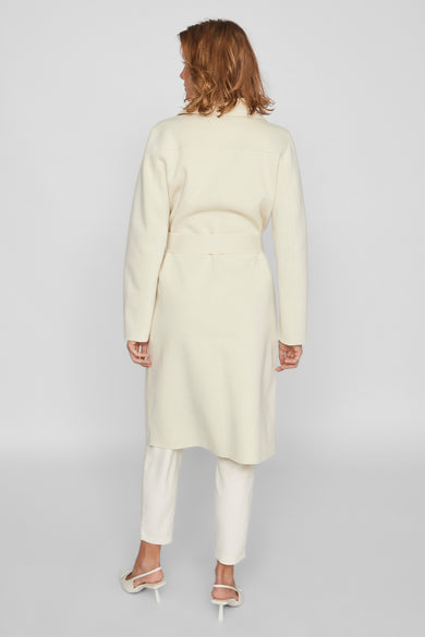 Ecru lange jas van VILA, met een ceintuur, gecombineerd met witte broek en hakken, van achteren gezien.
