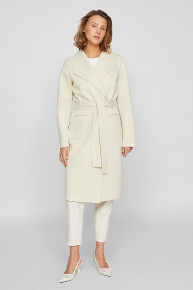 Ecru jas van VILA®, met ceintuur en zakken, gedragen met een witte broek en hakken.
