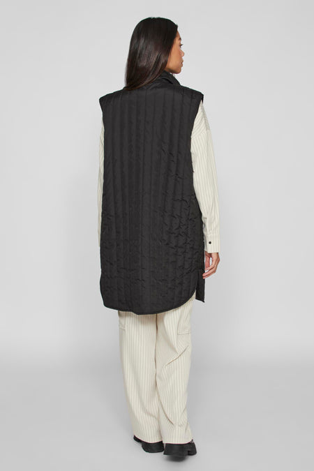 Zwarte bodywarmer van VILA, met verticale stiksels, gedragen over een gestreept overhemd en broek.