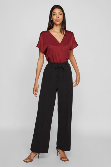 Zwarte broek van VILA®, met wijde pijpen en een strikceintuur, gecombineerd met bruine hakken en een bordeauxrode top.
