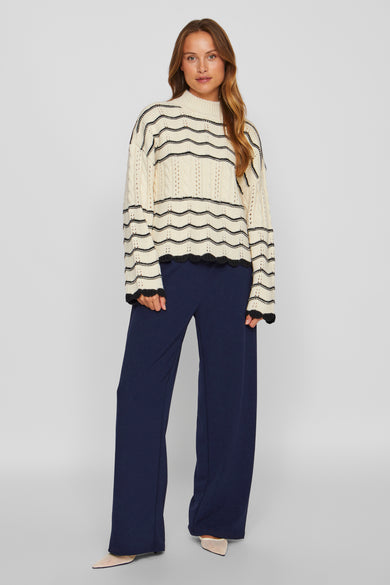 Gilet à col rond beige de VILA avec des rayures noires et des manches longues, associé à un pantalon bleu marine.