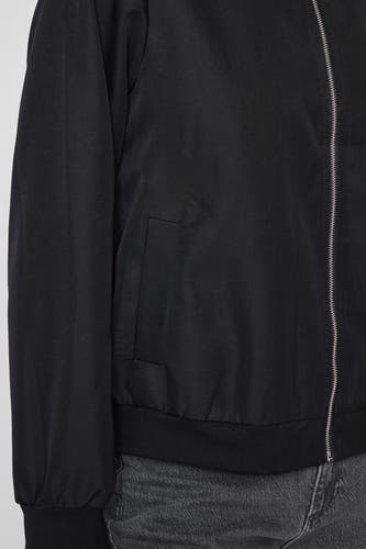 Manteau court noir de VILA, avec fermeture à glissière et poche poitrine, porté sur un jean gris.