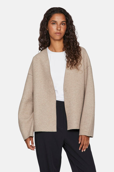 Beige korte jas van VILA, met lange mouwen en knoopsluiting, gedragen over een wit shirt en zwarte broek.

