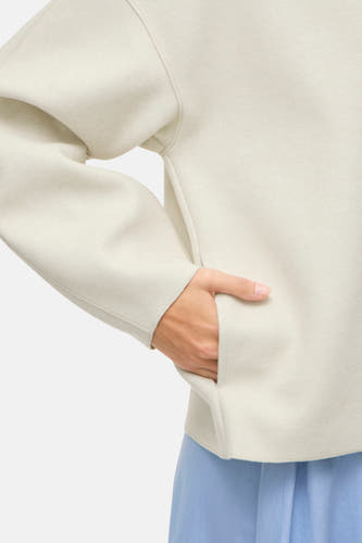 Ecru manteau court de VILA®, avec une main dans la poche et une jupe bleue en dessous.
