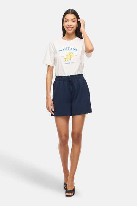 Donkerblauwe short van VILA® met hoge elastische taille en trekkoord, van gestructureerde stof. Gedragen met wit T-shirt en zwarte sandalen.