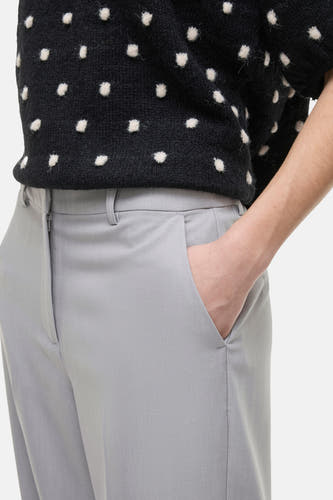 Grijze broek van VILA, met een hand in de zak, gecombineerd met een zwart gebreid shirt met witte stippen.

