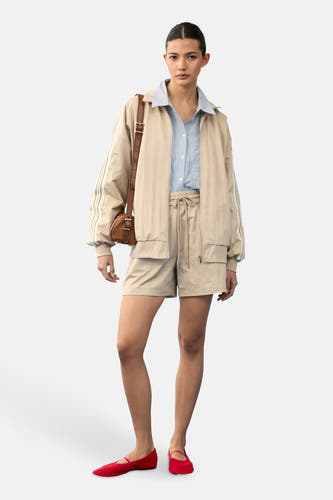 Beige short van VILA, met trekkoord en zakken, gecombineerd met een lichtblauw overhemd, jas en rode ballerina's.
