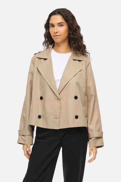 Manteau court beige de VILA® avec double boutonnage, manches longues et porté avec un t-shirt blanc et un jean noir.