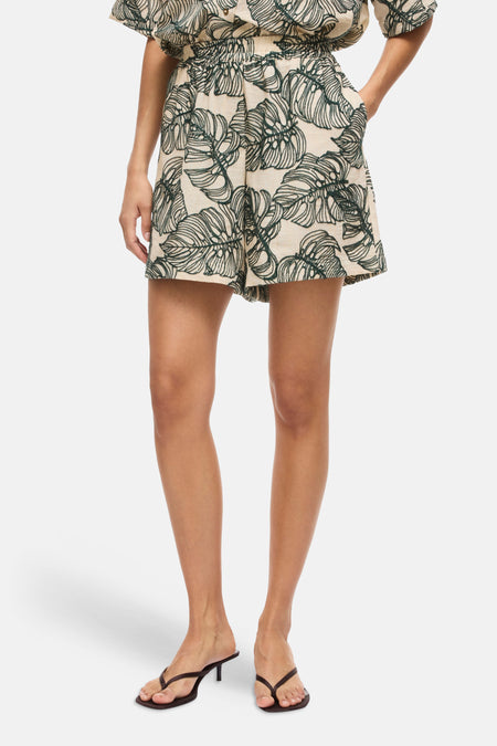 Short marron clair à motif de feuilles vertes de VILA, avec manches courtes, col et taille élastique, porté avec des sandales noires.