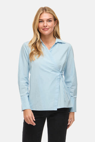 Chemise bleue à manches longues de VILA®, avec détail portefeuille sur le côté, portée avec un pantalon noir et des talons.