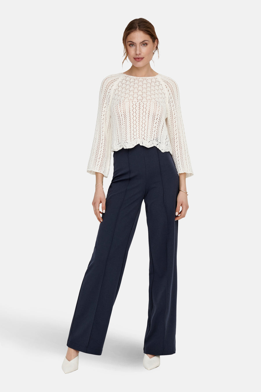 Ecru trui met ronde hals van ONLY, met ajourpatroon en driekwart mouwen, gecombineerd met een donkerblauwe pantalon en witte schoenen.
