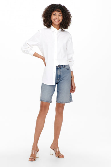 Korte bermuda van lichtblauw denim van ONLY, gecombineerd met een wit overhemd en transparante hakken.
