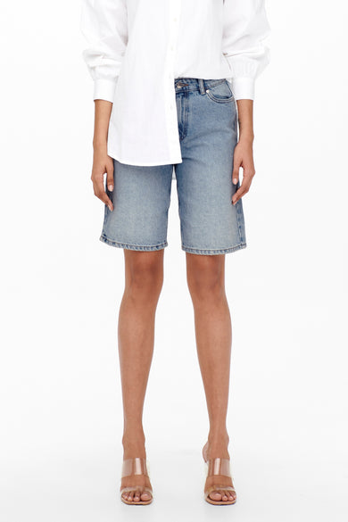 Korte, lichtblauwe denim bermuda van ONLY, gedragen met een wit overhemd en transparante hakken.
