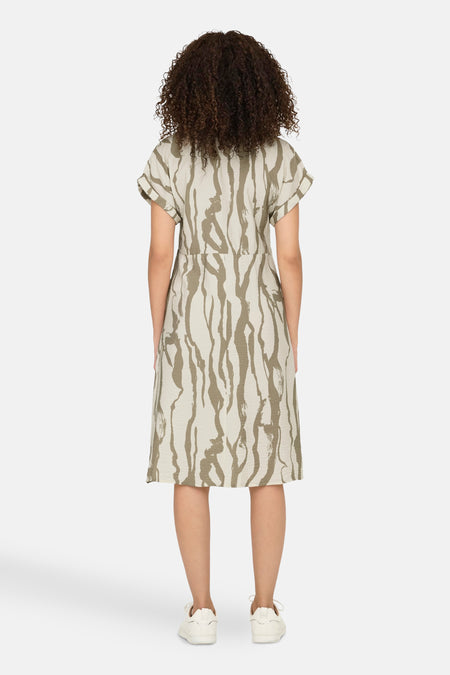 Vrouw met krullend haar van achteren, jurk met beige en olijfgroen patroon, witte sneakers, witte achtergrond.