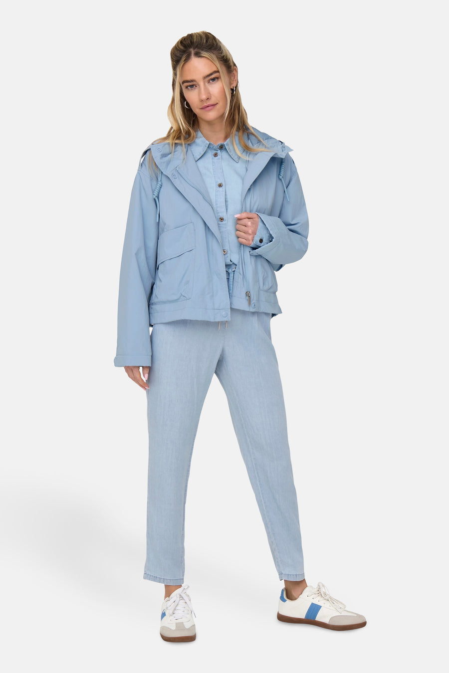 Vrouw draagt lichtblauwe outfit met jas, blouse, broek en witte sneakers.