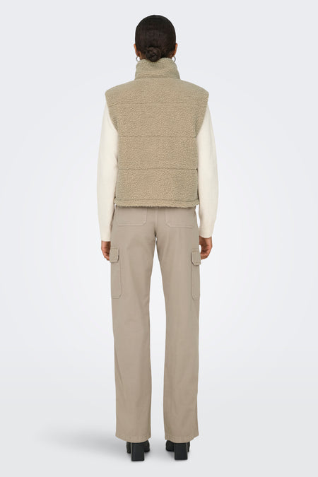 Doudoune sans manches en peau de mouton beige de ONLY®, dos avec col montant et coutures brodées, portée sur un pull blanc et un pantalon cargo beige.