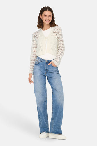 Vrouw draagt blauwe jeans, wit shirt en gehaakt vest, staand op witte achtergrond.