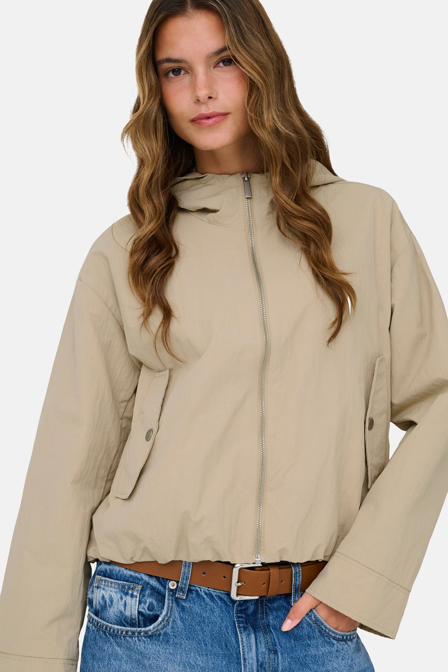 Beige jas van ONLY® met lange mouwen, ritssluiting en een bruine riem over een jeans.
