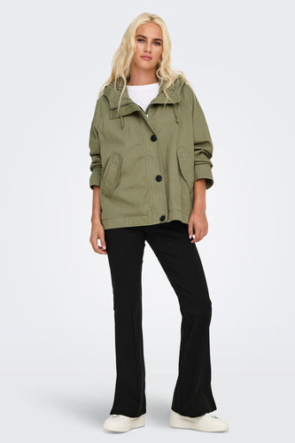 Vrouw in kaki jas en zwarte broek, staand voor witte achtergrond.