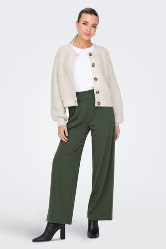 Femme en pantalon vert, chemise blanche et gilet beige avec des escarpins noirs.