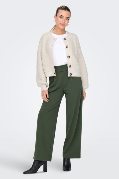 Femme en pantalon vert, chemise blanche et gilet beige avec des escarpins noirs.