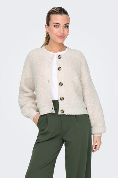 Veste écru de JDY, avec des manches longues, une fermeture à boutons et portée sur un chemisier blanc et un pantalon vert.