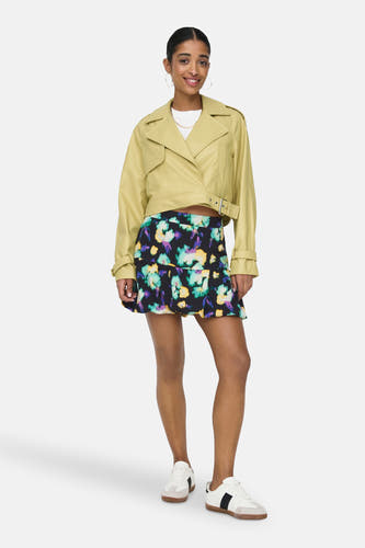 Gele jas van leer van ONLY, met lange mouwen, gedragen met een bloemenrok en witte sneakers.
