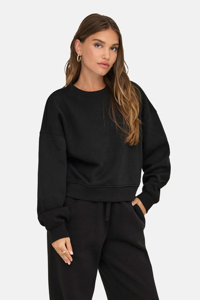 Zwarte sweater met ronde hals van ONLY, gedragen met een zwarte joggingbroek en gouden oorbellen.
