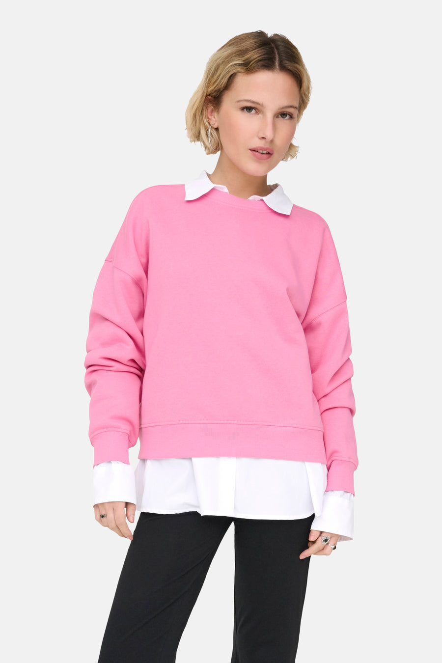 Vrouw in roze trui met witte blouse en zwarte broek, staand tegen een witte achtergrond.