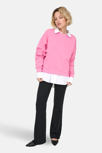 Vrouw draagt roze trui, witte blouse en zwarte broek staand.