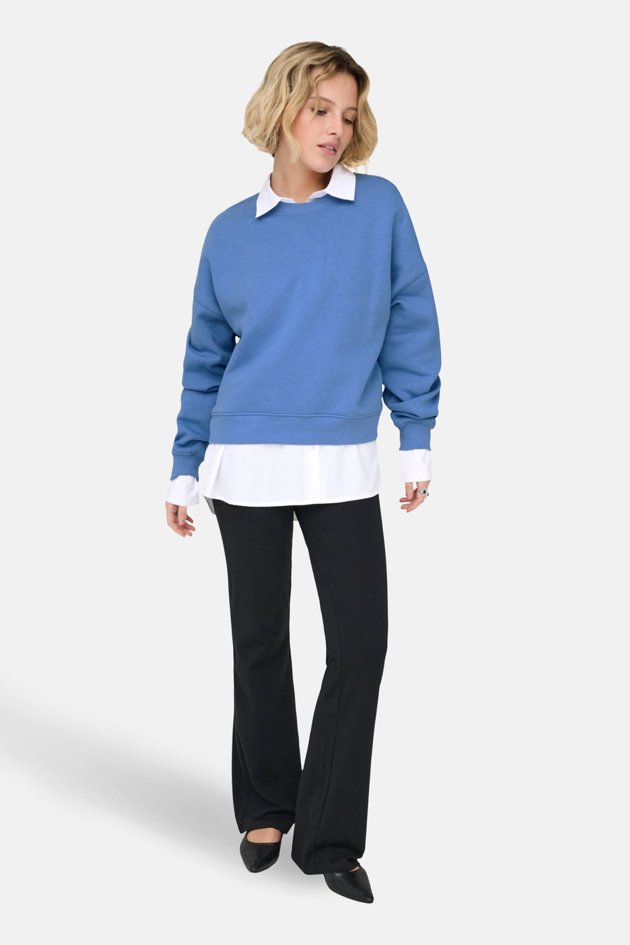 Vrouw in blauwe sweater, witte blouse, zwarte broek en schoenen, staand.