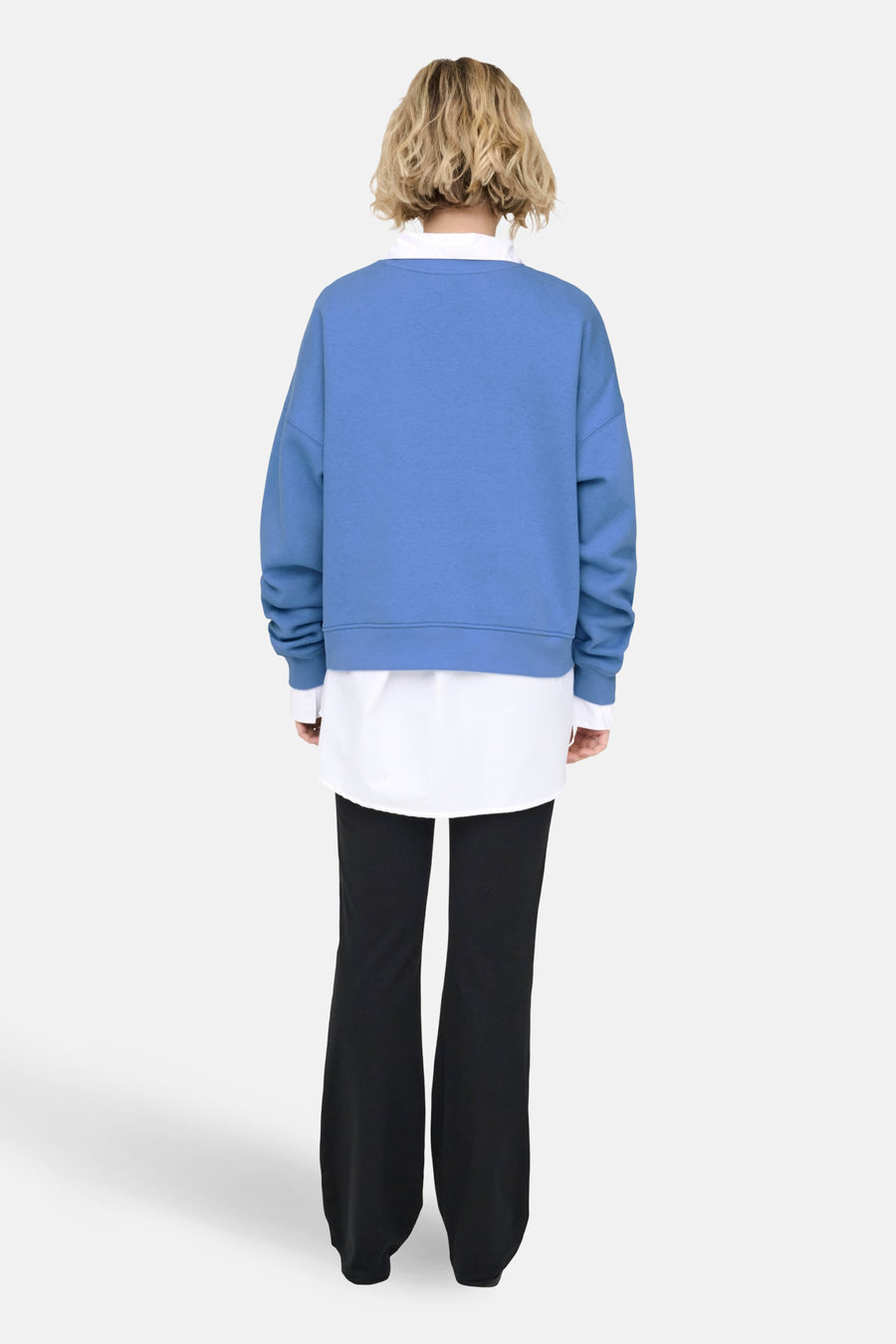 Achterkant van een vrouw in een blauwe trui, witte blouse en zwarte broek.